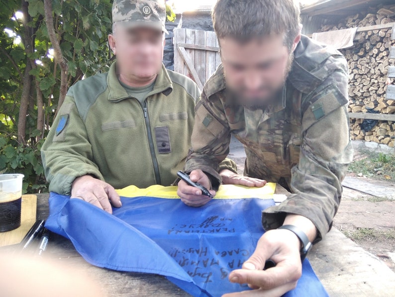 May include: Two people in military uniforms are signing a blue and yellow Ukrainian flag with black markers. The flag is laid out on a wooden table.