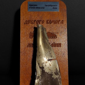 Könnte beinhalten: Ein Stück Metallpanzerung von einem russischen Fahrzeug, ausgestellt auf einer Holztafel mit dem Text "ДОКТОРО КЕШОРА" und "Originalteil der russischen Fahrzeugpanzerung Vom Schlachtfeld in der Ukraine".