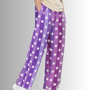 Puede incluir: Pantalones de pijama de pernera ancha con un estampado de rayas verticales en tonos morados y blancos. Las rayas se intercalan con formas de estrellas blancas. Los pantalones tienen bolsillos y son de tejido suave.