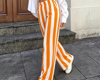Puede incluir: Pantalones de talle alto con un llamativo patrón de rayas verticales en naranja vibrante y blanco nítido. Estos pantalones de pierna ancha tienen una cintura elástica y un corte relajado y fluido que llega hasta los tobillos, combinados con zapatillas blancas.