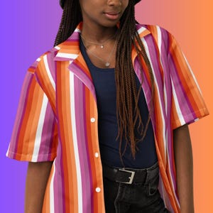 May include: Short-sleeved button-down shirt with vertical stripes in shades of orange, pink, and white. The shirt has a classic collar and is worn over a navy blue top. The model is wearing a black belt and dark jeans.