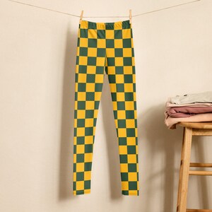 May include: Leggings featuring a yellow and dark green chequered design, hanging from a washing line. The garment is secured with a wooden peg. A small wooden stool and folded clothes are in the background.