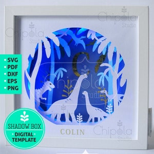 May include: A white shadow box with a blue and white paper cut design of a dinosaur in a forest. The box is lit from behind with LED lights. The name "COLIN" is written below the design. The box is labeled "SHADOW BOX - DIGITAL TEMPLATE" and "Chippola Studio".
