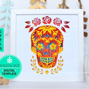 May include: A white frame containing a red, yellow and blue paper cut-out of a sugar skull with floral details. The skull is wearing a flower crown. The frame is on a white surface.