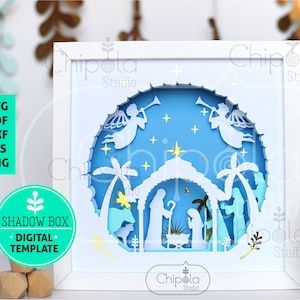 May include: A white shadow box with a blue and white paper cut design of the nativity scene. The design includes angels, palm trees, a star, and a manger with baby Jesus, Mary, and Joseph. The shadow box is framed in white and has a white background.