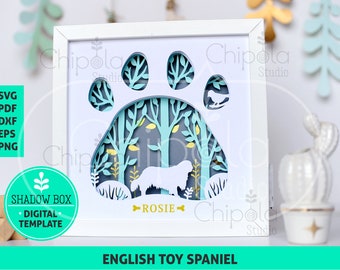 Puede incluir: Una caja de sombra blanca con un diseño de huella de pata. La huella de pata está recortada y el fondo es una escena de bosque azul y verde. El nombre "Rosie" está escrito en dorado en la parte inferior de la caja. El texto "ENGLISH TOY SPANIEL" está escrito en negro en la parte inferior de la imagen.