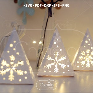 May include: Three white paper Christmas tree luminaries with cut-out snowflake designs. The trees are illuminated from within, creating a warm glow. The trees are arranged on a white surface.