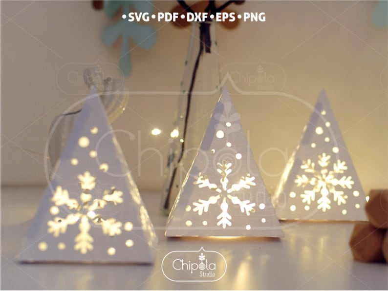 May include: Three white paper Christmas tree luminaries with cut-out snowflake designs. The trees are illuminated from within, creating a warm glow. The trees are arranged on a white surface.