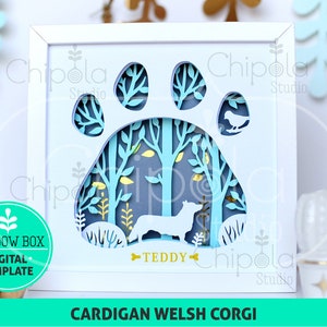 May include: A white shadow box with a blue paw print design. The paw print is filled with a forest scene with trees, leaves, and a small bird. A silhouette of a Cardigan Welsh Corgi dog is in the center of the paw print. The text "TEDDY" is below the dog.