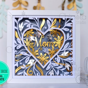 Könnte beinhalten: Eine weiße Schattenbox mit blauem Hintergrund und einem goldenen Herzen mit dem Wort "Mama" darin. Das Herz ist von goldenen und silbernen Blumen und Blättern umgeben.
