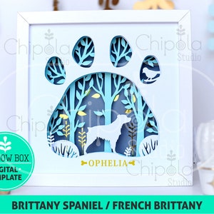 May include: A white frame with a blue and yellow paper cut-out design of a dog paw print with a forest scene inside. The dog is a Brittany Spaniel and the name "Ophelia" is written below the paw print. The text "Brittany Spaniel / French Brittany" is at the bottom of the frame.