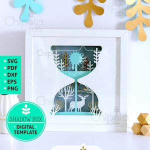 May include: A white frame with a blue and white paper cut-out design of a deer in a forest. The design is in the shape of an hourglass. The frame is on a white surface with a green circle that says "Shadow Box - Digital Template".