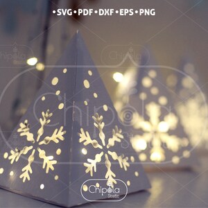 May include: White paper pyramid with a snowflake design and a light inside. The pyramid is decorated with white dots to resemble snow.