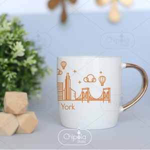 May include: White ceramic mug with a gold handle featuring a line drawing of the New York City skyline, including the Brooklyn Bridge, and the word "York" printed below the image.