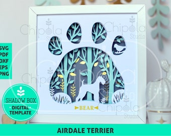 Puede incluir: Una caja de sombras enmarcada con un diseño de Airedale Terrier. La obra de arte incluye una huella de pata con una escena de bosque y una silueta de perro. La paleta de colores incluye blanco, verde azulado, dorado y gris. El texto "BEAR" está debajo.
