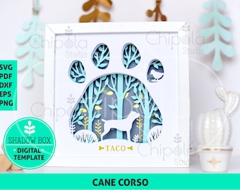 May include: A white shadow box with a paw print design. The paw print is cut out and the inside is decorated with a blue and green forest scene. A dog silhouette is in the center of the scene. The text "TACO" is below the dog. The text "CANE CORSO" is at the bottom of the box.