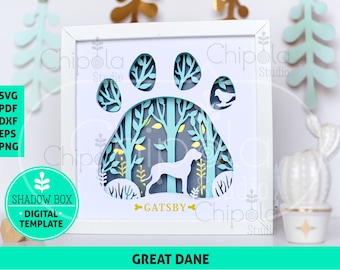 Puede incluir: Una caja de sombra blanca con un diseño de huella de pata azul. La huella de pata está llena de una escena de bosque con árboles, hojas y un perro. El perro es un Gran Danés llamado Gatsby. El texto "GREAT DANE" está en la parte inferior de la caja.