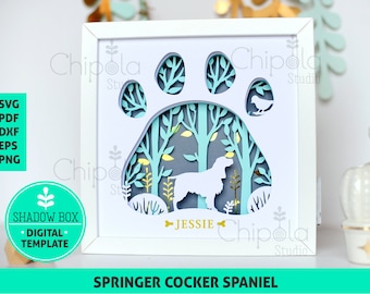Puede incluir: Un marco blanco con un diseño de huella de pata azul claro. Dentro de la huella de pata hay una escena de bosque con un perro blanco y el nombre "Jessie". El texto "SPRINGER COCKER SPANIEL" está en la parte inferior del marco.