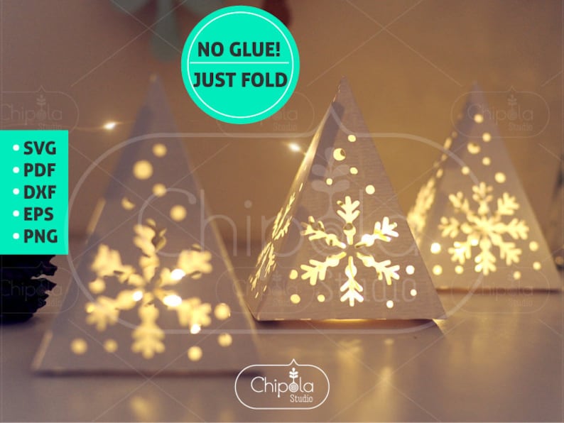 May include: Three white paper Christmas tree lanterns with snowflake cutouts and a string of lights inside. The lanterns are on a table and the text "NO GLUE! JUST FOLD" is in a green circle above them. The text "SVG, PDF, DXF, EPS, PNG" is listed on the left side of the image.