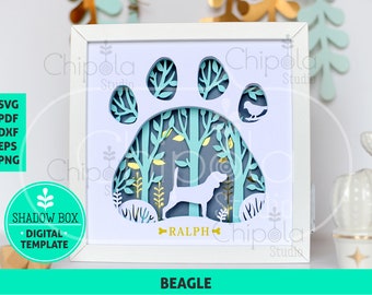 Puede incluir: Una caja de sombra blanca con un diseño de huella de pata. La huella de la pata está recortada y el interior está decorado con una escena de bosque y una silueta de perro beagle. El perro se llama "Ralph".