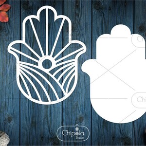 Peut inclure: Deux découpes de main de hamsa blanches sur un fond de bois bleu. La hamsa de gauche a un design détaillé avec des lignes et un cercle au centre. La hamsa de droite est un simple contour. Le texte "Chip" est visible sur la hamsa de droite.