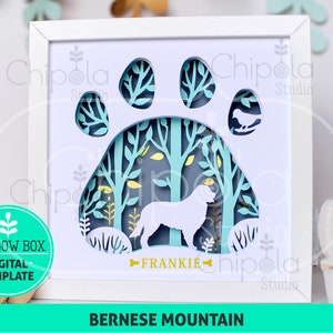 May include: A white shadow box with a blue paw print design. The paw print has a forest scene inside with a white silhouette of a Bernese Mountain Dog. The dog's name "FRANKIE" is written below the dog.