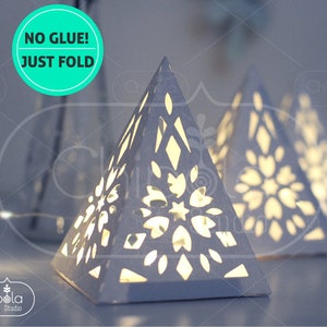 May include: Three white paper pyramid lanterns with intricate cut-out designs. The lanterns are illuminated from within, casting a warm glow. The text "NO GLUE! JUST FOLD" is visible in the top left corner of the image.