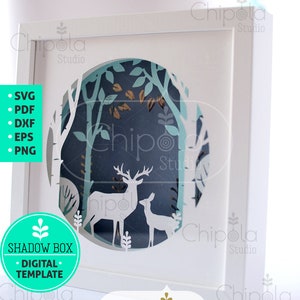 Peut inclure: Une boîte à ombre blanche avec une scène de forêt bleue et verte à l'intérieur. La scène représente deux cerfs, l'un debout et l'autre couché. La boîte est étiquetée "Shadow Box - Digital Template" et "Chipola Studio".
