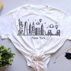 May include: A white t-shirt with a black outline of the New York City skyline, including the Statue of Liberty, the Empire State Building, and the Brooklyn Bridge. The text "New York" is printed below the skyline.