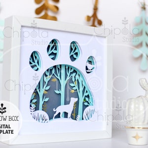 May include: A white shadow box with a paw print design. The paw print is cut out and the inside is decorated with a forest scene with a dog and birds. The text "SHADOW BOX DIGITAL TEMPLATE" is on the bottom left corner of the image.