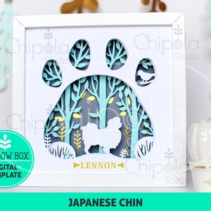 Op de afbeelding: Een digitaal sjabloon voor een schaduwdoos met een Japanse Chin-hond die in een bos scène staat. De hond is wit en het bos is blauw en groen. Het sjabloon bevat de naam van de hond, "Lennon", en de woorden "Japanse Chin".