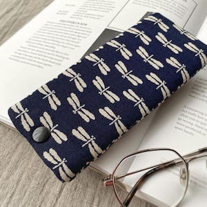 May include: A navy blue glasses case with a white dragonfly pattern. The case has a button closure and is lying on a book with a pair of glasses next to it.