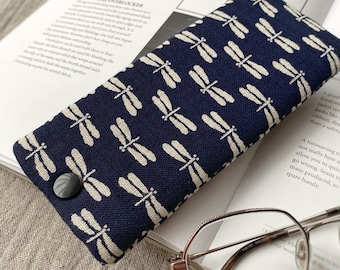 May include: A navy blue glasses case with a white dragonfly pattern. The case has a button closure and is lying on a book with a pair of glasses next to it.