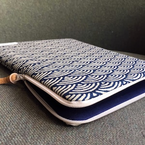 Könnte beinhalten: Eine Laptop-Hülle mit blau-weißem Muster, Reißverschluss und braunem Lederriemen. Die Hülle ist mit dunkelblauem Stoff gefüttert.