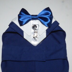 May include: A dark blue pet tuxedo garment featuring a white shirt collar, a shiny royal blue satin bow tie, and a central vertical white placket with two black buttons and a silver metal D-ring attachment for a leash or accessory.