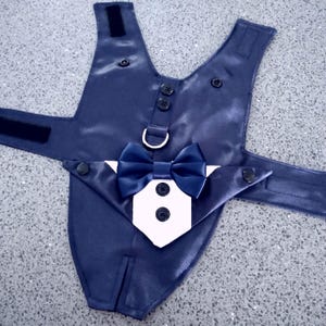 May include: A navy blue satin pet harness featuring a white shirt-front insert, a matching navy blue bow tie, two black buttons on the shirt front, two black buttons on the upper chest, and a silver D-ring. The harness includes black velcro straps.