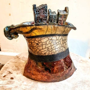May include: A ceramic vase with a unique, abstract design. The vase is shaped like a tilted, open container with a handle. The top of the vase features a row of small, textured houses. The vase is decorated with a crackled glaze in shades of brown, yellow, and black.