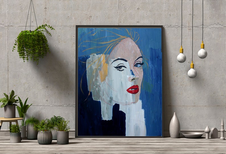 May include: Abstract painting of a woman's face with blue, white, and gold paint. The woman has red lips and blue eyes. The painting is framed in black.