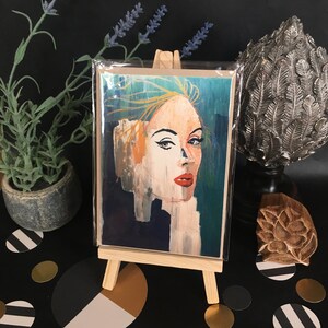 May include: A colorful abstract painting of a woman's face with blue, green, yellow, and red tones. The painting is on a card and displayed on a small easel.