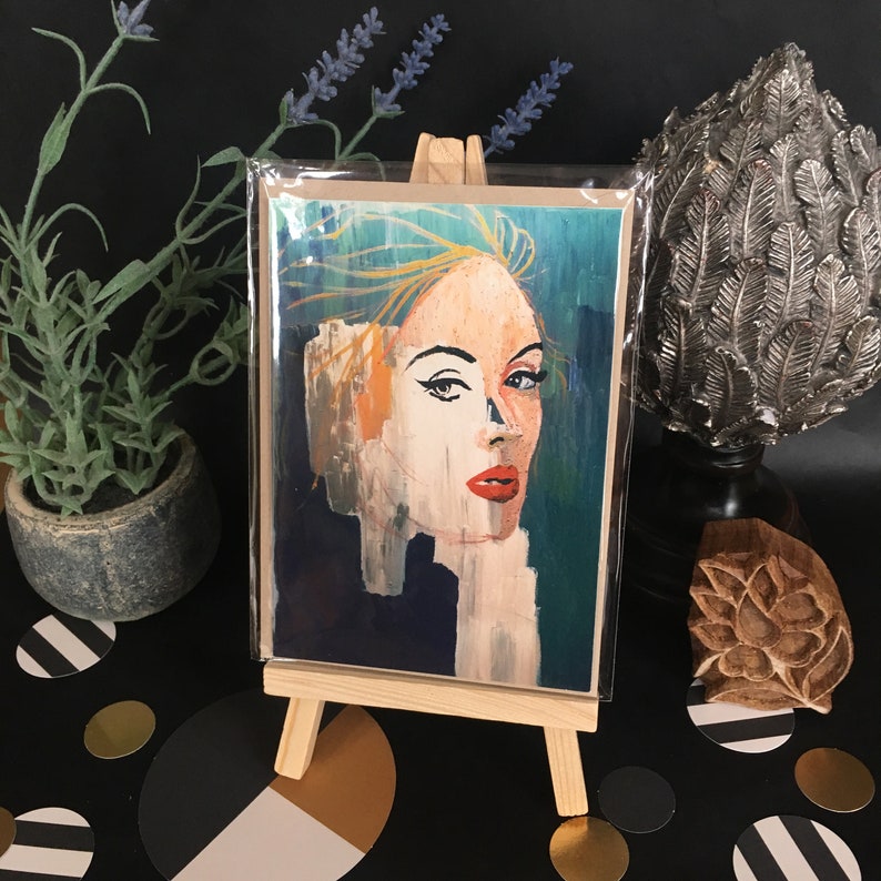 May include: A colorful abstract painting of a woman's face with blue, green, yellow, and red tones. The painting is on a card and displayed on a small easel.