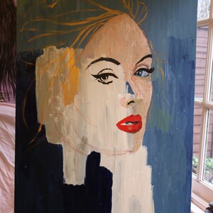 May include: A painting of a woman's face with a blue background. The woman has blonde hair and is wearing red lipstick. The painting is on a wooden easel.