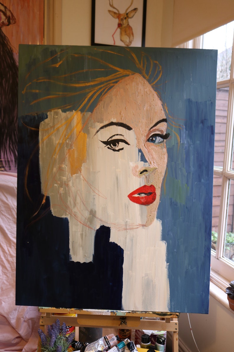 May include: A painting of a woman's face with a blue background. The woman has blonde hair and is wearing red lipstick. The painting is on a wooden easel.