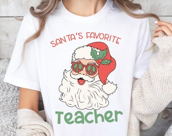 Puede incluir: Camiseta blanca con un gráfico de Santa Claus con gafas de sol y un signo de paz. El texto "Santa's Favorite Teacher" está impreso debajo del gráfico.
