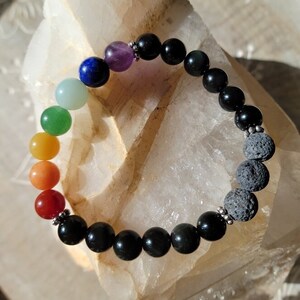 May include: A beaded bracelet featuring a spectrum of colors, transitioning from red to violet, with black obsidian and gray lava stone beads. The bracelet is strung on a stretch cord and accented with silver-colored metal beads.
