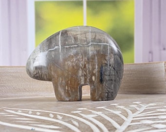 May include: A carved stone bear figurine in shades of gray and brown. The bear has a rounded body and a simple, stylized design. It is displayed on a wooden tray with a leaf pattern, with a window in the background.