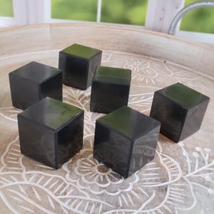 May include: Six dark gray, cube-shaped objects are arranged on a light-colored, carved wooden tray. The cubes have a smooth, reflective surface and are evenly spaced. The tray has a floral design.