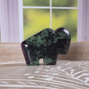 May include: A small, carved stone bison figurine. The bison is primarily black with green marbling, and has a smooth, polished finish. The figurine is standing on a light-colored wooden surface.