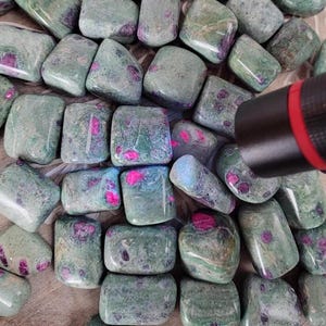 May include: A collection of tumbled stones, predominantly green with vibrant pink and purple inclusions. The stones have a smooth, polished appearance, and the arrangement suggests a natural, earthy aesthetic. The image is well-lit, highlighting the colors and textures of the stones.