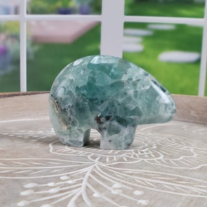 May include: A carved bear figurine made of blue-green fluorite crystal. The bear is standing on a decorative wooden tray. The crystal has a translucent quality, with visible internal patterns and inclusions. The background shows a window with a view of a green yard.