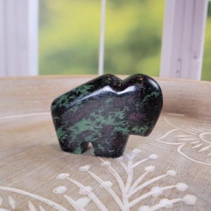 May include: A small, polished stone carving of a bison. The sculpture is primarily black with green and maroon veining. The bison is standing on a light-colored wooden surface with a floral design. The background is a blurred view of a window.